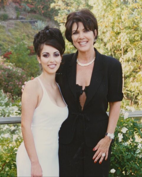 Kris Jenner i dorosła Kim Kardashian pozują razem na zewnątrz, otoczone zielenią i krzewami. Kris stoi po prawej stronie, w czarnym garniturze z krótkimi rękawami i naszyjnikiem z pereł, obejmując ramieniem Kim. Kim stoi po lewej stronie w białej sukience na ramiączkach, z ciemnym upięciem włosów i mocniejszym makijażem.