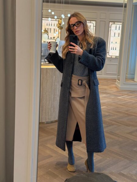 Kobieta w lustrze, ubrana w długi, szary wełniany płaszcz, szary sweter z warkoczowym wzorem i beżową spódnicę o kopertowym kroju z szerokim paskiem. Na nogach ma botki na obcasie w połączeniu z długimi, szarymi skarpetami. Trzyma kubek z napojem i telefon, robiąc selfie. Ten zestaw to inspiracja, jak nosić spódnice zimą.