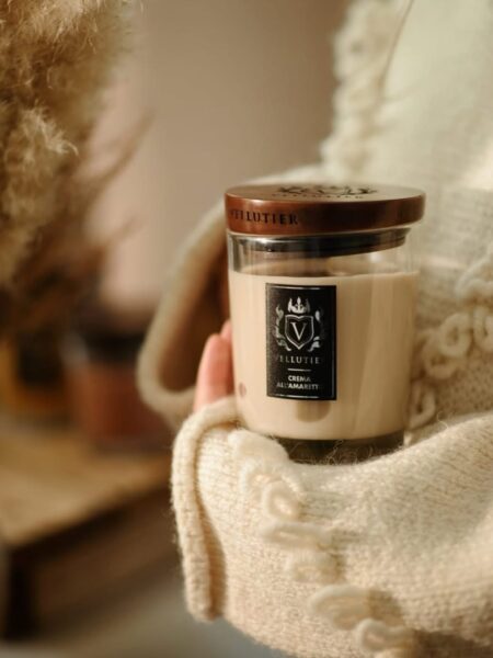 Ujęcie z bliska na dłonie ubrane w gruby, kremowy, wełniany sweter, które trzymają średnią jesienną świecę zapachową marki Vellutier. Świeca o beżowym wosku i zapachu 'Crema All'Amaretto' ma drewnianą przykrywkę. Scena jest ciepła i przytulna, z miękkim, rozproszonym światłem w tle, symbolizująca domowy relaks i luksus.