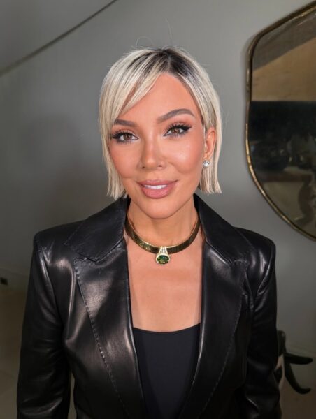Ujęcie w półpostaci przedstawia kobietę (prawdopodobnie Kris Jenner w peruce) o jasnych, platynowych włosach z ciemnymi odrostami, ściętych w krótkiego boba. Ma na sobie czarną, skórzaną marynarkę/żakiet i czarny top. Nosi gruby, złoty naszyjnik typu żmijka z dużym, zielonym kamieniem (szmaragdem). Uśmiecha się, ma mocny makijaż oczu i małe kolczyki. W tle widoczne jest lustro w złotej ramie.