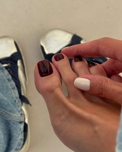 Zbliżenie na stopy z modnym pedicurem hybrydowym. Paznokcie pomalowane są głębokim, lśniącym kolorem burgundowym/ciemnym bordo, z wyjątkiem jednego paznokcia (drugi palec) pomalowanego jasnym, kremowym, niemalże białym lakierem jako akcent. Na zdjęciu widoczna jest również dłoń delikatnie dotykająca stopy oraz fragmenty niebieskich jeansów i granatowo-białych, znoszonych trampek. To jesienna/zimowa inspiracja na stylowy pedicure burgundowy z białym paznokciem.