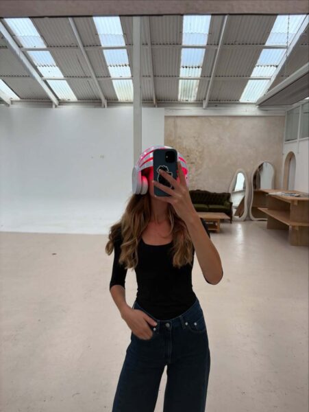 Kobieta robi sobie selfie w dużym, jasnym studiu. Ma na sobie czarny top z rękawami 3/4 i ciemne jeansy. Jej twarz jest zasłonięta smartfonem, który trzyma. Na głowie ma urządzenie w kształcie hełmu lub słuchawek, które wygląda jak do terapii czerwonym światłem na włosy lub terapia LED na włosy, emitujące czerwone i białe światło. Ma długie, falowane, brązowe włosy. W tle widać duże lustro, sofę, drewniany stół i wysoki, biały sufit z oknami.