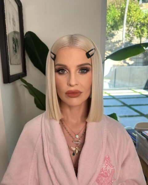 Portret kobiety z jasnym, prostym bobem i czarnymi spinkami, ubranej w różowy szlafrok. Jej twarz jest mocno konturowana, z wyraźnie zaznaczoną linią żuchwy i zapadniętymi policzkami. Taki wygląd często określa się terminem "twarz ozempikowa".