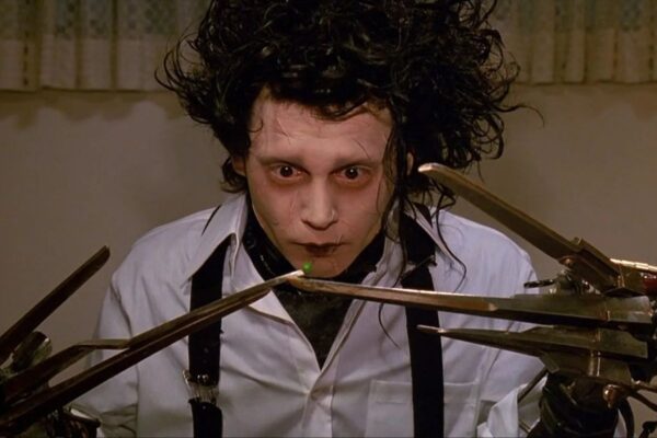 Kadr z filmu przedstawiający postać o bladym makijażu, z dzikimi włosami i nożycami zamiast dłoni, wpatrującą się w zielony groszek trzymany między ostrzami.