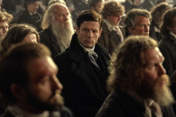 Mężczyzna w eleganckim czarnym stroju siedzi wśród tłumu podczas debaty politycznej. Scena z serialu „Ród Guinnessów”.