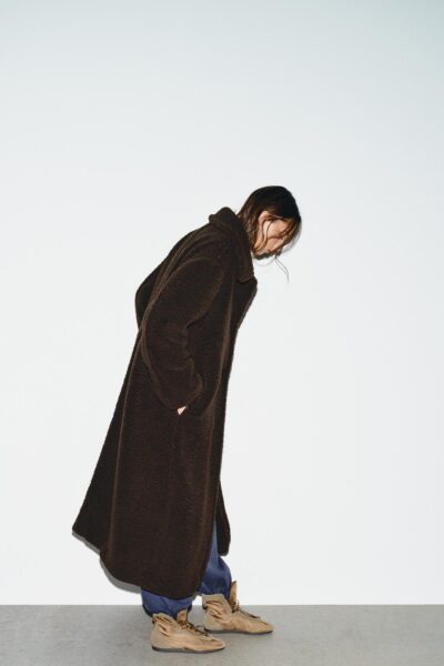 Modelka w długim, brązowym płaszczu typu teddy coat wykonanym z miękkiego materiału o fakturze misia. Stylizacja utrzymana w minimalistycznym tonie – oversize’owy fason, proste linie i casualowe buty w odcieniu karmelu. Ujęcie studyjne na białym tle podkreśla rzeźbiarską formę sylwetki i teksturę tkaniny.