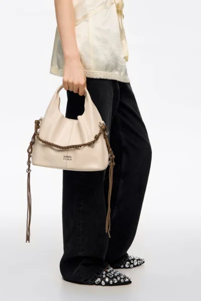 Zbliżenie na beżową torebkę Bimba Y Lola trzymaną w dłoni modelki. Torebka ma miękką, marszczoną formę, ozdobną brązową plecioną rączkę z metalowymi elementami i długie skórzane troczki. Stylizacja dopełniona jest czarnymi denimowymi spodniami i czubkami butów udekorowanych dużymi kryształami.