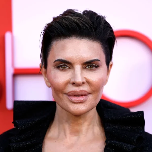 Lisa Rinna – zbliżenie twarzy aktorki w krótkiej fryzurze o zaczesanych do tyłu włosach, w czarnym żakiecie z wysokim kołnierzem. Naturalny makijaż z mocno podkreślonymi ustami i brwiami, neutralne tło w odcieniu różu i czerwieni.