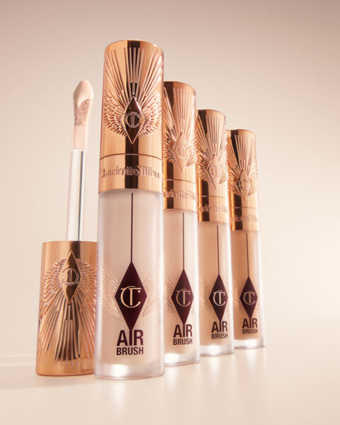 Zbliżenie na korektor Airbrush marki Charlotte Tilbury w eleganckim, matowym opakowaniu z metaliczną zakrętką w kolorze różowego złota. Luksusowa kompozycja czterech buteleczek ustawionych w rzędzie na beżowym tle podkreśla premium charakter produktu.