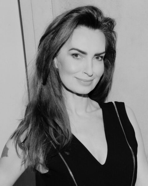 Zdjęcie czarno-białe: portret uśmiechniętej kobiety z długimi, ciemnymi włosami, ubranej w elegancką, czarną sukienkę z zamkiem błyskawicznym. Agnieszka Maciąg przyczyna śmierci, ikona stylu i autorka poradników.