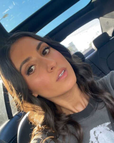 Zbliżenie na twarz kobiety z długimi, ciemnymi włosami, ujęte w samochodzie z otwartym szyberdachem. Ma na sobie szary, sprany T-shirt z grafiką. Makijaż jest naturalny, podkreślający urodę: świetliste, brązowe smoky eyes, zmysłowe usta z błyszczykiem i wyraźnie zaznaczony róż/bronzer na policzkach, dający efekt "słonecznego" blasku.