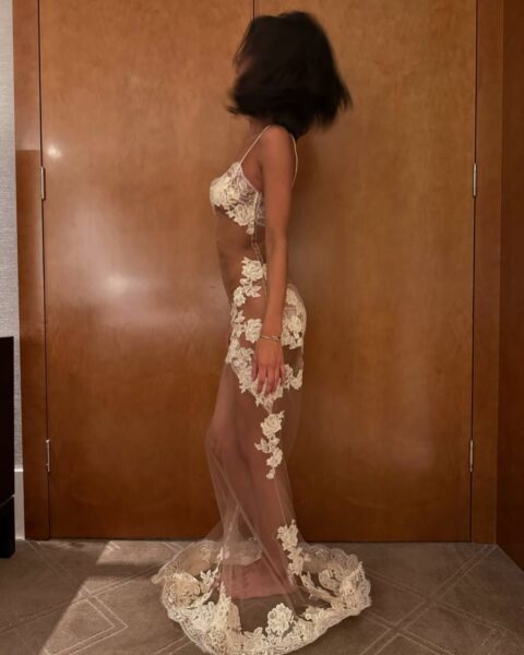 Kobieta, Angelina Censori, uchwycona w dynamicznej pozie z rozmyciem ruchu włosów, stojąca bokiem do kamery przed drewnianymi drzwiami. Ma na sobie przejrzystą, długą suknię lub narzutkę z delikatnego tiulu lub siateczki, ozdobioną białymi, koronkowymi aplikacjami w kształcie kwiatów i liści. Pod spodem widać beżową lub cielistą bieliznę. Na nadgarstku nosi delikatną bransoletkę. Długie, prześwitujące elementy sukni układają się wokół jej stóp.