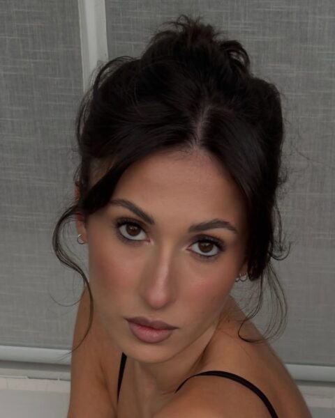 Zbliżenie na twarz kobiety, Angelina Censori, patrzącej prosto w obiektyw, z ujęciem od góry. Ma ciemne włosy upięte w luźny kok z wyciągniętymi pasmami wokół twarzy. Ubrana jest w czarny top na cienkich ramiączkach. Makijaż jest naturalny, ale wyrazisty: wydłużone rzęsy, subtelne brązowe cienie, podkreślony bronzerem kontur twarzy oraz usta w kolorze nude. W tle widać białe lub jasnoszare, tekstylne rolety.