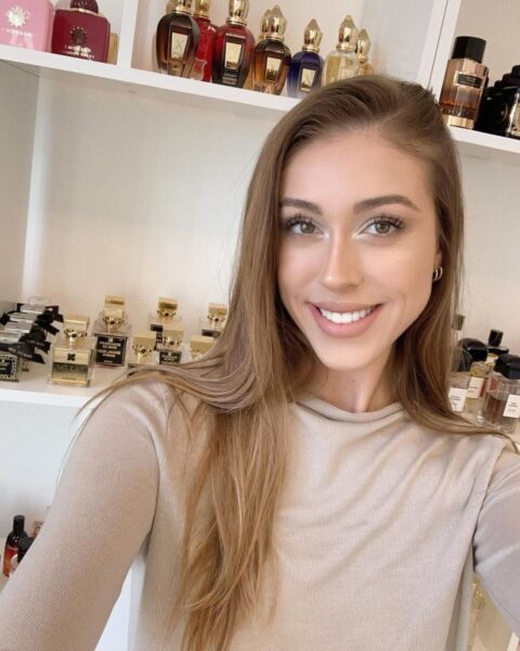 Zbliżenie na uśmiechniętą, elegancką kobietę z długimi, jasnobrązowymi włosami i naturalnym makijażem, ubraną w beżową bluzkę. W tle, na białych półkach, widoczna jest duża kolekcja luksusowych i niszowych flakonów perfum, w tym marki Amouage i Xerjoff. Zdjęcie typu "selfie" oddaje radość z posiadania i używania pięknych zapachów. Hashtagi: ulubione perfumy niszowe, kobiecy uśmiech, minimalistyczny makijaż.