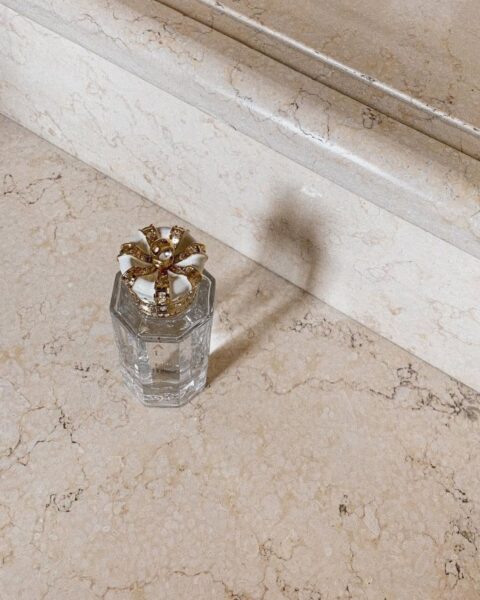 Elegancki flakon perfum o geometrycznym kształcie z ozdobnym, złotym korkiem w kształcie rozety, umieszczony na tle marmurowych schodów w ciepłym, beżowym odcieniu. Kompozycja zdjęcia podkreśla luksusowy i estetyczny charakter produktu, sugerując ekskluzywność i dbałość o detale. Obraz symbolizuje luksus, wyrafinowanie i unikalność.