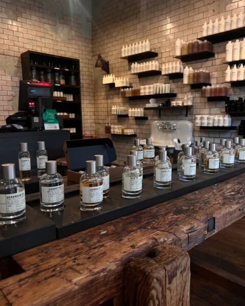 Wnętrze nowoczesnego butiku z niszowymi perfumami (Le Labo), utrzymane w stylu industrialnym. Na pierwszym planie widoczne są flakony perfum o minimalistycznym designie (m.in. Geranium 33, Ambrette 9, Jasmin 17) ustawione na długiej, masywnej, drewnianej ladzie. Tło stanowi ściana z białej cegły, na której wiszą półki z licznymi butelkami kosmetyków do ciała. Zdjęcie oddaje atmosferę poszukiwania kultowych, minimalistycznych zapachów i luksusowego rzemiosła.