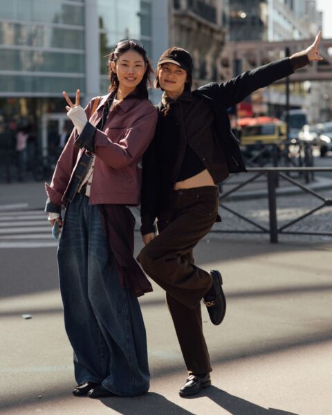 Dwie uśmiechnięte, młode kobiety w modnych, luźnych stylizacjach street style, pozujące na słonecznej ulicy. Pierwsza, po lewej, ma na sobie bardzo szerokie, workowate jeansy, biały top i oversizową, różowo-fioletową kurtkę, z przewiązaną w pasie koszulą w kratę. Druga, po prawej, ma na sobie czarną kurtkę, brązowe, proste spodnie i czapkę z daszkiem, a jej poza jest bardziej dynamiczna. Obie prezentują trend na swobodne, warstwowe i miejskie sylwetki.