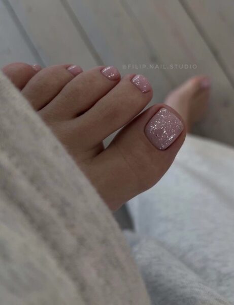 Pedicure w pastelowym różu z połyskiem – paznokcie pokryte delikatnym brokatowym lakierem tworzą efekt subtelnego blasku. Stylizacja minimalistyczna i kobieca, idealna do casualowych stylizacji.