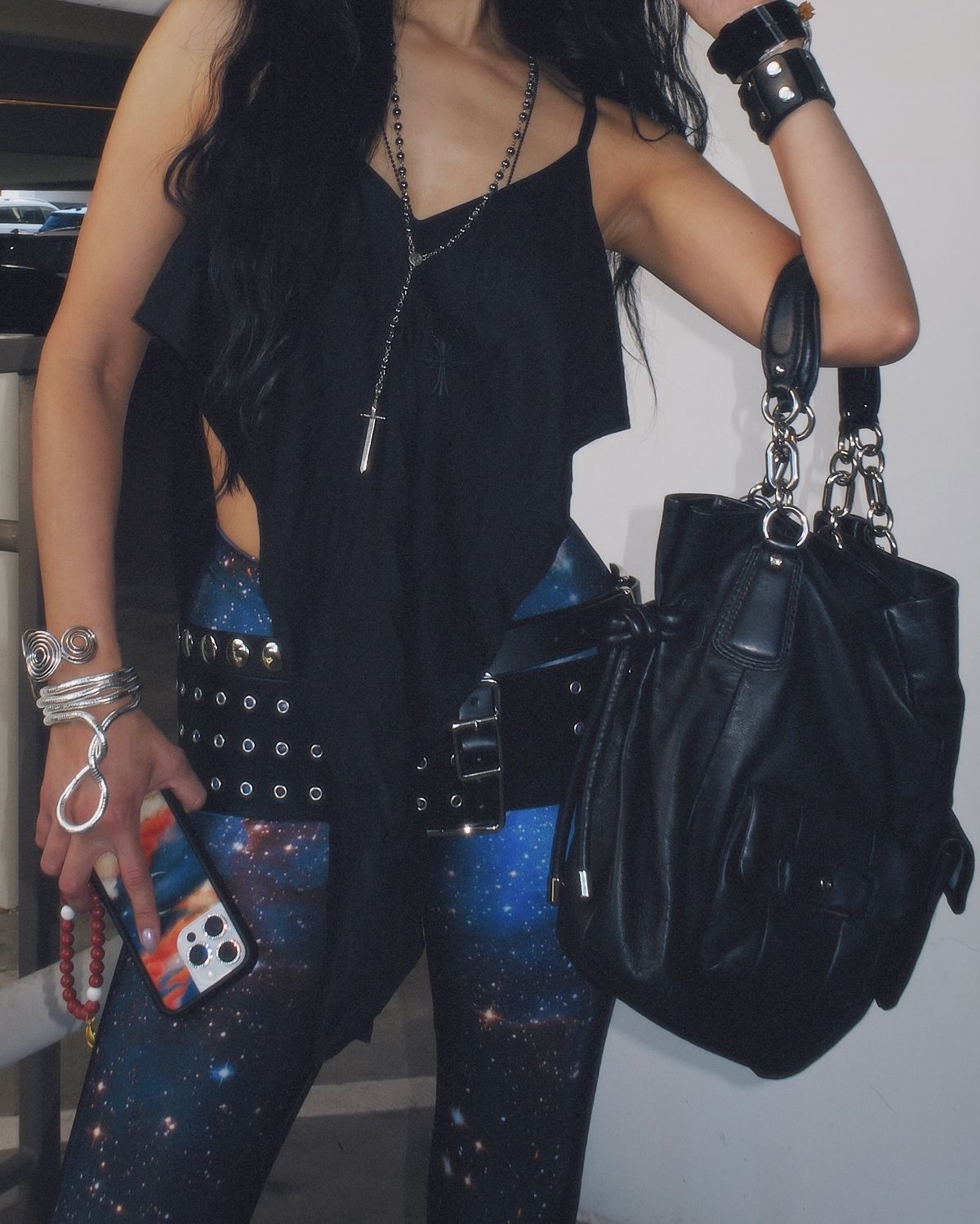Kosmiczny wzór trend: Stylizacja w duchu cyber-grunge – kobieta w czarnym topie na ramiączkach z surowym wykończeniem, galaktycznych legginsach z nadrukiem gwiazd, szerokim pasie z metalowymi oczkami i masywnych bransoletkach. W dłoni trzyma telefon w etui w kolorach kosmicznego nieba i czarną torbę z grubymi łańcuchami.