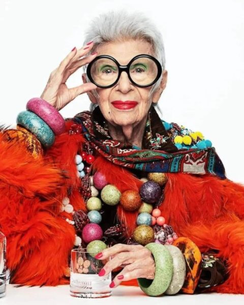 Iris Apfel w jaskrawopomarańczowym futrze, z okrągłymi okularami, mocnym makijażem ust i dużą ilością kolorowej, warstwowej biżuterii.