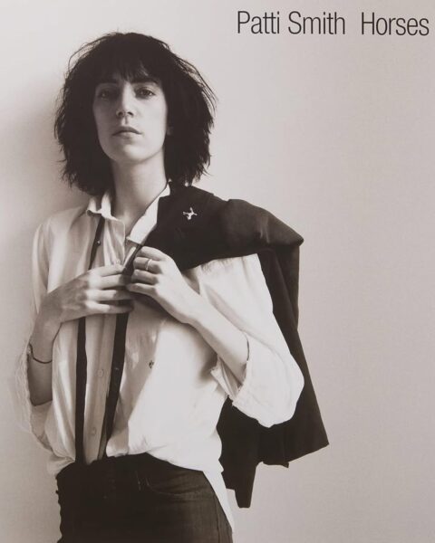 Patti Smith, czarno-białe zdjęcie. Ma rozpiętą białą koszulę, na ramieniu przerzuconą marynarkę i krawat. Tytuł: Patti Smith Horses.