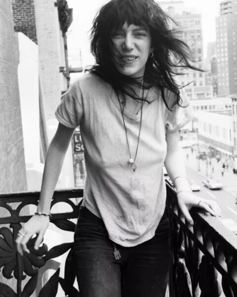 Patti Smith na balkonie hotelu Chelsea. Czarno-białe zdjęcie, luźny T-shirt, naszyjniki i rozwiane włosy.