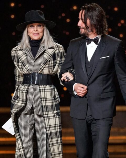 Diane Keaton w kraciastej stylizacji, z szerokim czarnym paskiem, kapeluszem i golfem, prowadzona przez Keanu Reevesa w smokingu.