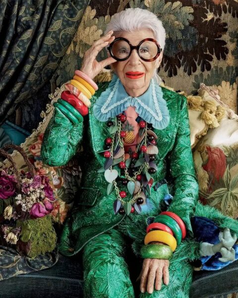 Iris Apfel w zielonym garniturze w deseń, z falbankowym kołnierzykiem, wielkimi okularami i mnóstwem kolorowej biżuterii.