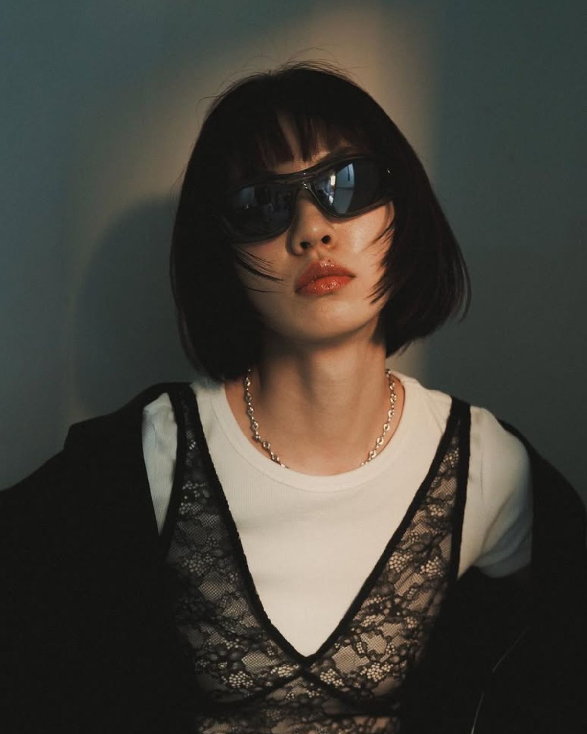 Zdjęcie modowe przedstawiające kobietę z krótkimi, ciemnymi włosami w stylu messy bob, ubraną w czarny, koronkowy top na białej koszulce i czarną marynarkę nonszalancko zarzuconą na ramiona. Uzupełnieniem stylizacji są futurystyczne, czarne okulary przeciwsłoneczne i gruby, srebrny łańcuszek. Tło jest stonowane, a twarz kobiety jest oświetlona dramatycznym, bocznym światłem, podkreślającym jej makijaż w odcieniach nude. Inspiracja: jak Japonki pielęgnują włosy i tworzą swoje charakterystyczne stylizacje.