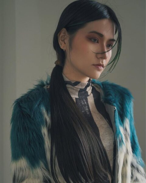 Modowe zbliżenie na kobietę w stylizacji z futerka (faux fur) w odcieniach turkusu, bieli i czerni, narzuconego na dopasowany golf z graficznym, czarno-beżowym wzorem. Włosy modelki są ciemne, długie i proste, zebrane w niski kucyk, a ich nieskazitelna gładkość zwraca uwagę. Makijaż jest wyrazisty, z akcentem na smokey eye w odcieniach brązu i czerwonawych różu. Scena oświetlona jest miękkim, rozproszonym światłem. Inspiracja dla fanek minimalizmu i luksusu w pielęgnacji: jak Japonki pielęgnują włosy, by osiągnąć efekt tafli.