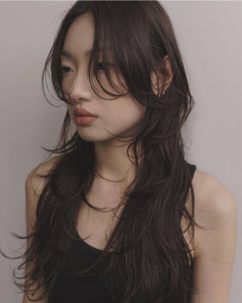 Zbliżenie na twarz i ramiona kobiety o długich, pofalowanych, ciemnobrązowych włosach. Fryzura jest modnym połączeniem warstwowego cięcia, tzw. wolf cut lub mullet, z grzywką typu curtain bangs, która miękko okala twarz. Makijaż utrzymany jest w ciepłych, matowych odcieniach brzoskwini i terakoty. Kobieta ubrana jest w prosty, czarny top na ramiączkach. Tło jest neutralne, jasnoszare, a oświetlenie studyjne podkreśla objętość i teksturę włosów, oddając inspirację japońskim stylem fryzur.