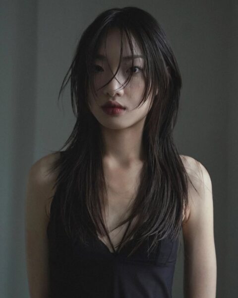 Portret modowy młodej kobiety z długimi, ciemnymi włosami o wyraźnej, warstwowej strukturze, przypominającej modny wolf cut lub shag. Część włosów opada naturalnie na twarz, tworząc nonszalancki, ale zmysłowy efekt. Modelka ubrana jest w prosty, czarny top na ramiączkach z dekoltem w serek. Makijaż jest subtelny, z akcentem na ciemniejszą, matową pomadkę w odcieniu wina lub bordo. Zdjęcie utrzymane jest w ciemnej, nastrojowej tonacji z miękkim, rozproszonym oświetleniem.