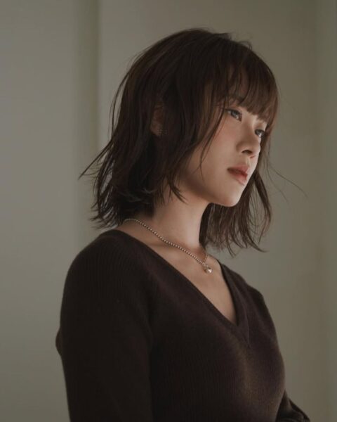 Portret kobiety z włosami o długości do podbródka, w modnym, postrzępionym cięciu typu shaggy bob, z lekką grzywką sięgającą brwi. Włosy mają głęboki, jesienny odcień ciemnego brązu. Modelka ubrana jest w brązowy sweter z dekoltem w serek i ma na sobie delikatny srebrny łańcuszek z wisiorkiem oraz liczne srebrne pierścionki. Stoi przy oknie z drewnianymi żaluzjami, patrząc w bok. Światło dzienne nadaje zdjęciu ciepły, jesienny klimat, podkreślając teksturę fryzury.