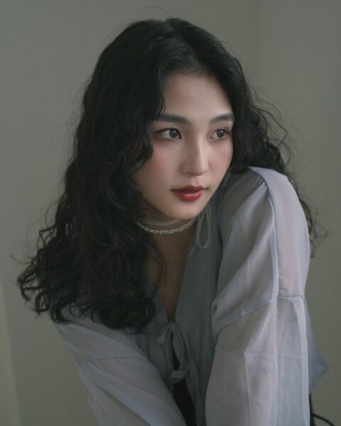 Portret ukazujący popiersie młodej kobiety z długimi, czarnymi, gęstymi włosami w intensywnych, drobnych lokach i falach, nadających im dużej objętości. Ubrana jest w zwiewną, jasnoszarą bluzkę z głębokim dekoltem w serek wiązanym pod szyją. Centralnym elementem stylizacji jest krótki naszyjnik typu choker z białych pereł. Makijaż podkreśla oczy i usta – na ustach widoczna jest matowa, ciemnoczerwona szminka. Zdjęcie utrzymane jest w romantycznym, nieco eterycznym klimacie z miękkim oświetleniem.