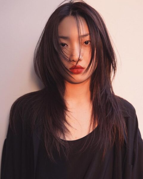 Intensywny portret modowy kobiety o długich, ciemnych, warstwowo ciętych włosach, które swobodnie opadają na twarz i ramiona. Ubrana jest w luźną, czarną koszulę lub top. Makijaż jest dramatyczny, z ustami pomalowanymi na głęboki, matowy odcień czerwieni, kontrastujący z prostą stylizacją. Scena jest oświetlona silnym, ciepłym, bocznym światłem (najprawdopodobniej zachodzącego słońca), które nadaje zdjęciu kinowy, mroczny klimat.