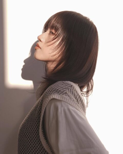 Ujęcie z profilu przedstawiające kobietę z krótkimi, ciemnymi włosami o geometrycznym, warstwowym cięciu na wysokości ramion i prostą grzywką. Włosy są gładkie i lśniące. Kobieta ubrana jest w jasną koszulę i dzianinową, szarą kamizelkę. Tło jest jasne, białe, a światło słoneczne, padające z boku, tworzy wyraźny, dramatyczny cień na ścianie i oświetla profil modelki. Na ustach delikatny, brzoskwiniowy makijaż.