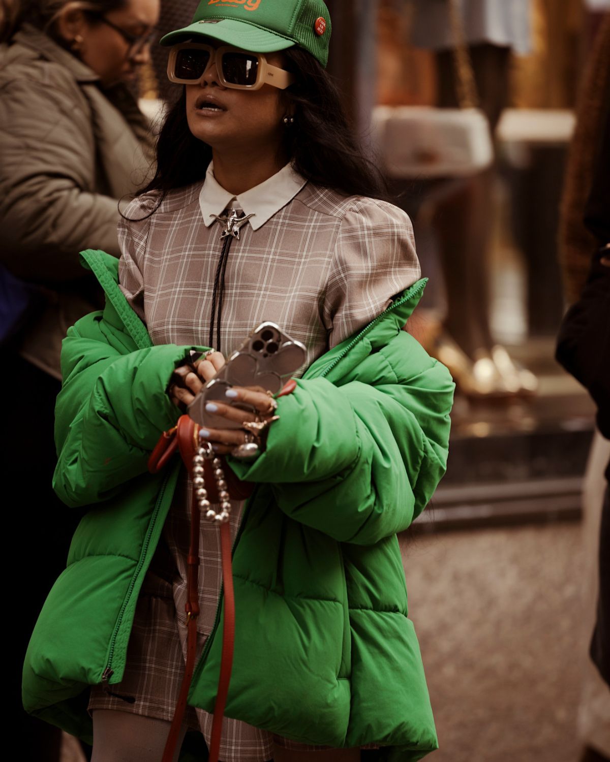 Kobieta w modnym, miejskim stroju trzyma w rękach telefon i zsuwa z ramion oversizową zieloną kurtkę puchową. Ma na sobie sukienkę w beżowo-brązową kratę z białym kołnierzykiem, ozdobioną naszyjnikiem bolo. Całość dopełniają duże, beżowe okulary przeciwsłoneczne i zielona czapka z daszkiem. Zdjęcie ilustruje, jak nosić kurtki puchowe w nowoczesnej i eleganckiej stylizacji, łącząc je z klasycznymi wzorami i oryginalnymi akcesoriami.