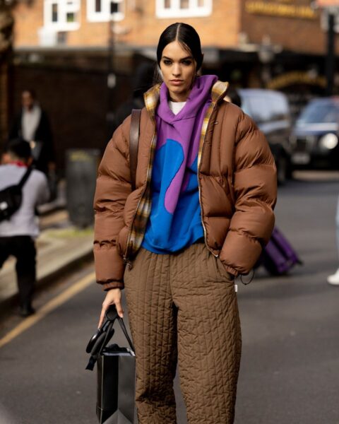 Modelka pozuje na ulicy w zimowej stylizacji. Ma na sobie brązową, pikowaną kurtkę puchową typu oversized, pod którą widać bluzę z kapturem w intensywnych kolorach (fioletowy i niebieski abstrakcyjny wzór) oraz biały podkoszulek. Dół stroju stanowią pikowane, brązowe spodnie o szerokiej nogawce, dopasowane do kurtki i białe, masywne buty. Kobieta trzyma w dłoniach czarne rękawiczki i małą papierową torbę. Zdjęcie przedstawia inspirację modą uliczną na zimę, łączącą wygodę i najnowsze trendy.