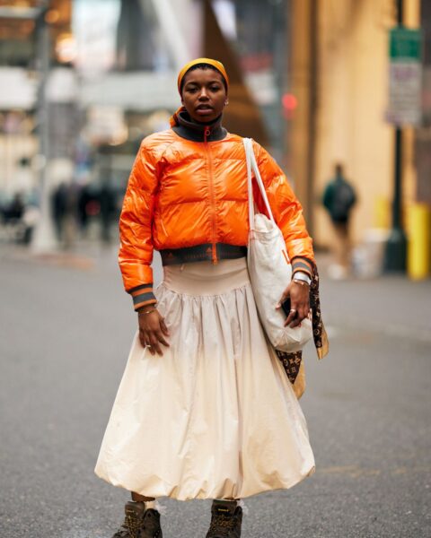Modna kobieta w zimowej stylizacji miejskiej. Ma na sobie krótki, błyszczący, pomarańczowy bomber z szarymi ściągaczami. Kurtka została zestawiona z długą, beżową spódnicą o bufiastym kroju, sięgającą do połowy łydki. Na głowie nosi żółtą opaskę lub turban, a całości dopełniają solidne, brązowe buty trekkingowe i duża, biała, materiałowa torba z wzorzystą apaszką. Zdjęcie doskonale ilustruje łączenie sportowej, funkcjonalnej odzieży z eleganckimi elementami, co jest kluczowe w trendach, które pokazują jak nosić kurtki puchowe w nieoczywisty sposób.