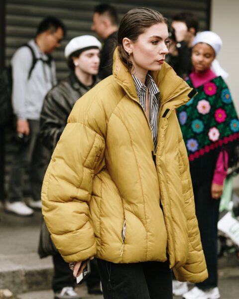 Stylowa kobieta idąca ulicą w modnej, zimowej stylizacji. Ma na sobie musztardową, oversizową kurtkę puchową z wysokim kołnierzem, a pod nią elegancką koszulę w czarno-białe paski. Dół stroju stanowią czarne dżinsy lub spodnie. Kobieta ma gładko zaczesane włosy i makijaż z podkreślonymi ustami. Zdjęcie doskonale demonstruje, jak nosić kurtki puchowe w eleganckim, a jednocześnie nowoczesnym wydaniu, łącząc je z klasycznymi elementami garderoby.