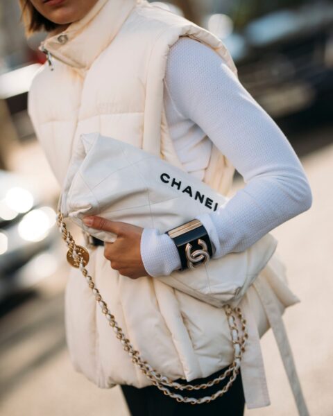 Ujęcie z bliska na stylową kobietę w eleganckiej, zimowej stylizacji. Ma na sobie kremową, pikowaną kamizelkę puchową z wysokim kołnierzem, założoną na biały, dopasowany golf z długim rękawem. W ręku trzyma dużą, pikowaną, kremową torebkę z czarnym napisem "CHANEL" i grubym złotym łańcuchem. Na nadgarstku widać szeroką, czarną bransoletę ze złotym logo. Zdjęcie doskonale demonstruje, jak nosić kurtki puchowe (w tym przypadku kamizelki) w luksusowej, monochromatycznej odsłonie, z wykorzystaniem akcesoriów od znanych projektantów.