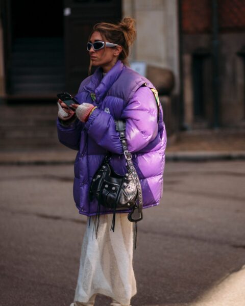 Modna kobieta w stroju ulicznym, pozująca na tle miejskiego krajobrazu. Ma na sobie błyszczącą, oversizową, fioletową kurtkę puchową z wysokim kołnierzem, zestawioną z długą, beżową spódnicą lub spodniami z dzianiny. Całość uzupełniają futurystyczne, wąskie okulary przeciwsłoneczne, czarna torebka-worek na ramieniu oraz beżowe, miękkie buty, przykryte luźnym materiałem. Zdjęcie ilustruje, jak nosić kurtki puchowe w odważnej kolorystyce i z modnym połączeniem kontrastujących faktur i fasonów.