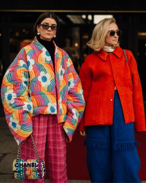 Dwie modnie ubrane kobiety stojące na czerwonym dywanie przed wejściem do budynku, prezentując odważne zimowe stylizacje. Po lewej stronie: kobieta w obszernej, pikowanej kurtce puchowej w tęczowy wzór z motywami kół i kwiatów. Ma na sobie różowe spodnie w kratę i małą torebkę Chanel. Po prawej stronie: kobieta w krótkiej, pomarańczowo-czerwonej kurtce z kołnierzykiem, zestawionej z szerokimi, niebieskimi spodniami z frędzlami. Obie noszą ciemne okulary przeciwsłoneczne. Zdjęcie ilustruje, jak nosić kurtki puchowe w kolorowym, radosnym i maksymalistycznym stylu.