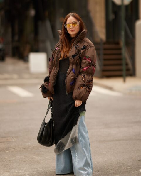 Kobieta w wyjątkowej stylizacji ulicznej, stojąca na skrzyżowaniu. Ma na sobie brązową, krótką kurtkę puchową ozdobioną kolorowymi, naszywanymi motylami. Pod spodem widać długą, czarną sukienkę, spod której wystają szerokie, błyszczące spodnie dżinsowe w jasnoniebieskim kolorze. Całość uzupełniają żółte okulary przeciwsłoneczne, czarna torebka na ramię i czerwone czubki butów. Zdjęcie inspiruje i pokazuje, jak nosić kurtki puchowe w stylu łączącym elegancję z odważnym, eklektycznym designem.