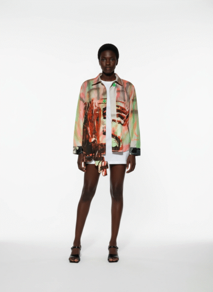 Kobieta pozuje na białym tle w artystycznej stylizacji — oversizowa koszula z abstrakcyjnym nadrukiem w odcieniach zieleni, czerwieni i czerni zestawiona z krótką, białą sukienką i czarnymi sandałkami. Całość utrzymana w duchu streetwearowej elegancji.