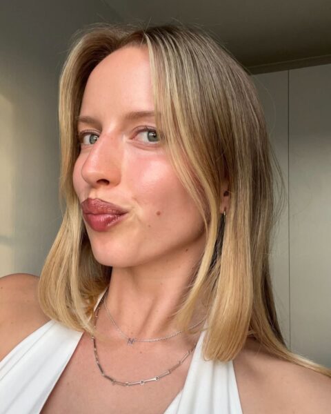 Portret młodej kobiety z jasnymi włosami i naturalnie podkreślonym makijażem, ubranej w biały top typu halter. Jej skóra promienieje zdrowym blaskiem, a usta są błyszczące. Patrzy bezpośrednio w obiektyw z figlarnie zmarszczonymi ustami. Na szyi ma delikatne srebrne naszyjniki. Zdjęcie doskonale oddaje efekt, jaki można uzyskać stosując kremy, które zastępują wizytę u kosmetyczki, podkreślając naturalne piękno i pielęgnację.