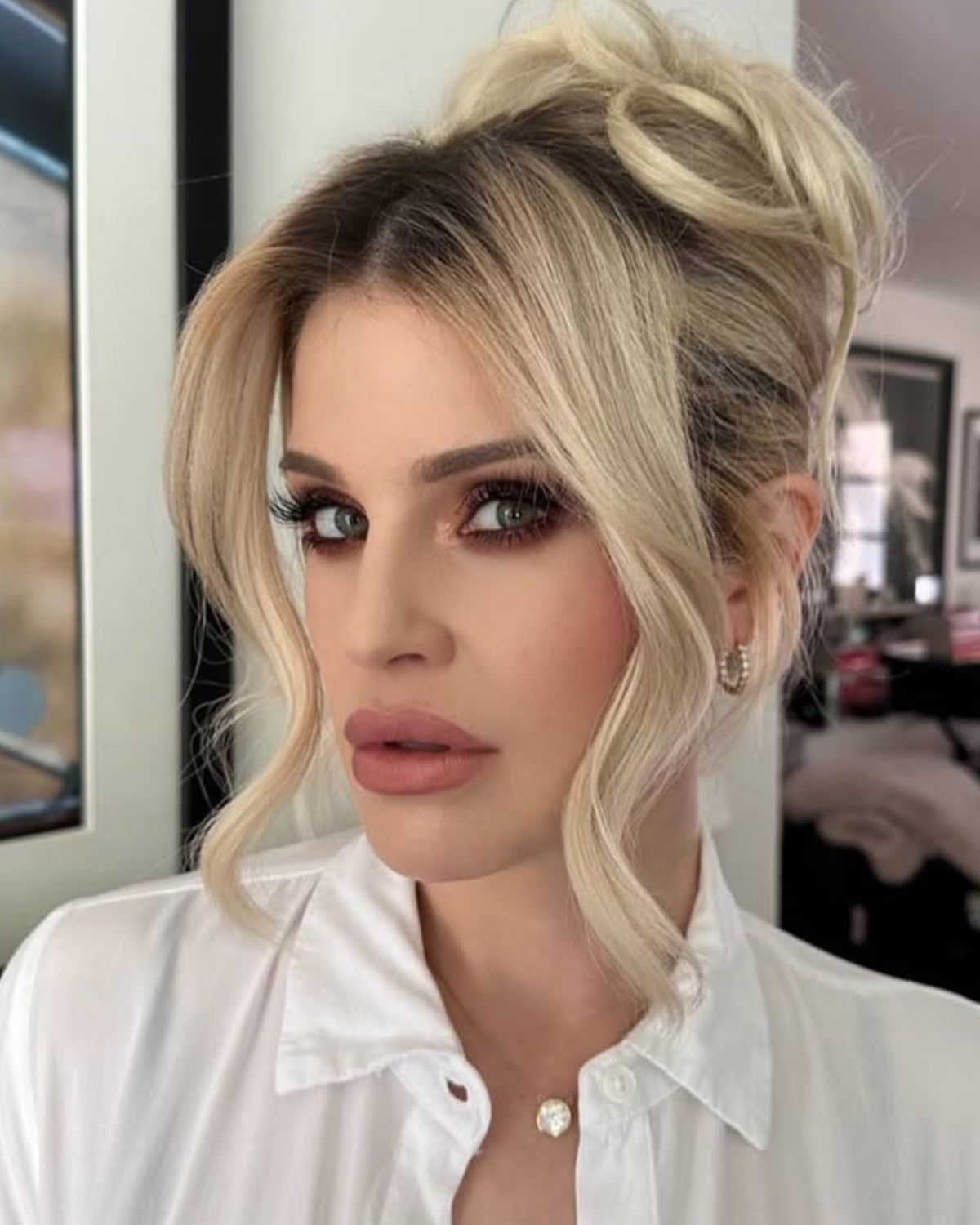 Blondynka w białej koszuli z kołnierzykiem i zmysłowym makijażem, z włosami upiętymi w wysoki, lekko niechlujny kok, patrzy prosto w obiektyw. Na szyi ma delikatny naszyjnik z zawieszką w kształcie kwiatu. Zdjęcie ma naturalne oświetlenie, w tle widoczny jest fragment wnętrza. Użyj kremy na twarz ozempikową dla kobiet z klasą.