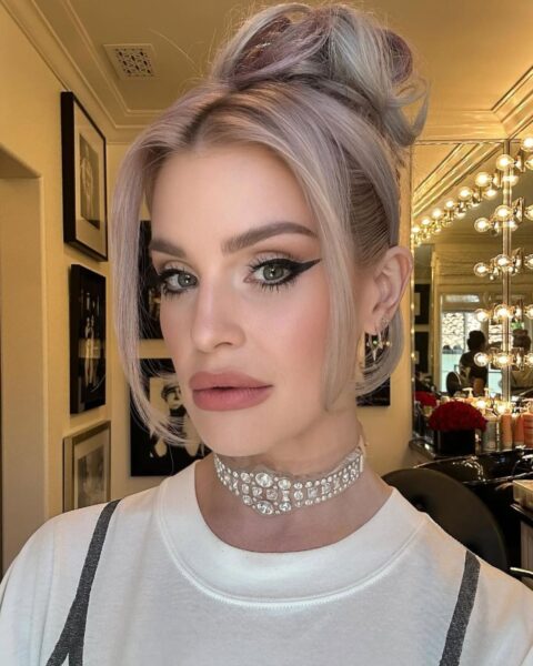 Kelly Osbourne w zbliżeniu na twarz, z włosami w kolorze lawendowo-blond upiętymi w wysoki, niedbały kok z luźnymi pasmami przy twarzy. Ma na sobie biały T-shirt z szarymi ramiączkami i szeroki, bogato zdobiony naszyjnik typu choker z kryształów. Makijaż oczu jest intensywny, z wyraźną kreską w stylu "kocie oko" i brzoskwiniowo-różowymi ustami. W tle widoczna jest toaletka z lustrem otoczonym żarówkami.