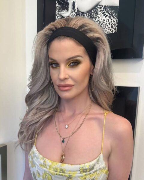 Kelly Osbourne w stylizacji inspirowanej stylem retro/lat 60., z długimi, blond włosami ułożonymi z dużą objętością, ozdobionymi czarną opaską. Ma na sobie letnią sukienkę na cienkich ramiączkach, w kolorze kremowo-żółtym z żakardowym, kwiatowym wzorem. Uwagę przykuwa wyrazisty makijaż oczu z neonowo-żółtymi cieniami. Na szyi ma kilka warstw złotych naszyjników. W tle wiszą czarno-białe zdjęcia w ramkach.