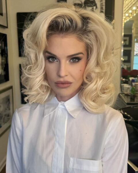 Blondynka w stylizacji inspirowanej Marylin Monroe, z jasnymi, platynowymi włosami ułożonymi w obszerne, Hollywoodzkie loki do ramion, z dużą objętością. Ma na sobie klasyczną, białą koszulę z kołnierzykiem, zapiętą pod szyję. Makijaż jest wyrazisty, z przydymionymi oczami (smoky eye) w szaro-brązowej tonacji i matowymi, neutralnymi ustami. Kobieta patrzy prosto w obiektyw. W tle widać ramy zdjęć i fragment salonu urody z lustrem.